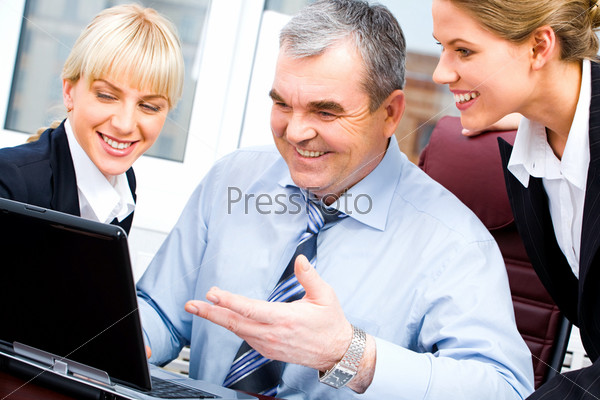 Portrait of senior boss and two young businesswomen looking at the laptop monitor with smiles on the background of office window