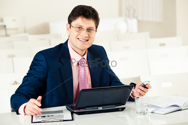 Photo of smiling businessman at work sitting by laptop and holding mobile phone