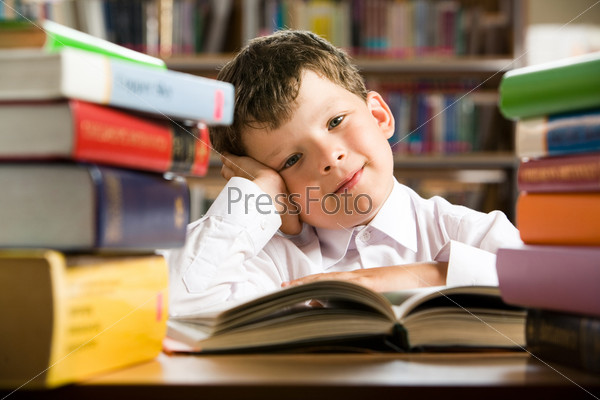 Portrait of cheerful lad sitting in library before book and looking at camera with smile