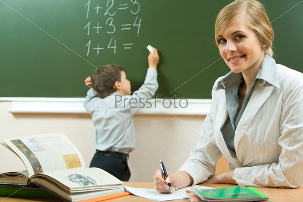 Portrait of pretty teacher sitting at the table on the background of schoolboy