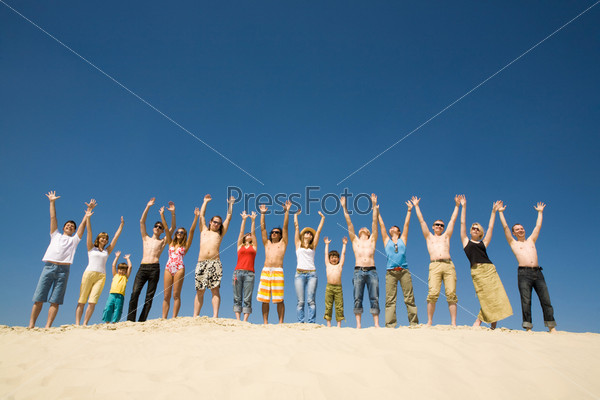 Image of many friends standing on sandy beach with their arms raised against blue sky