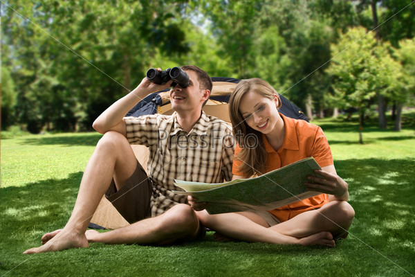 Photo of resting couple sitting on green grass while girl reading paper and guy watching through binoculars