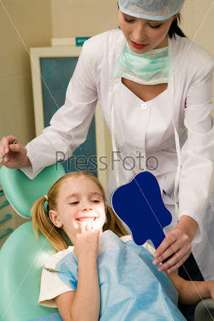 Photo of happy girl looking at her teeth in the mirror with helpful assistant near by
