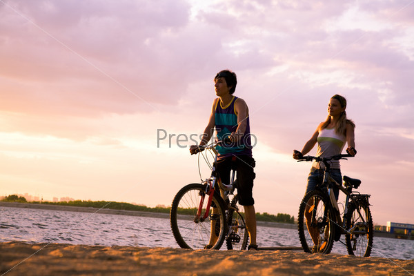 Figures of happy couple standing on seashore with their bicycles and enjoying sunset