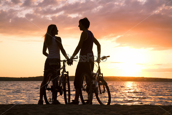 Figures of couple standing on seashore with their bicycles and looking at each other at sunset