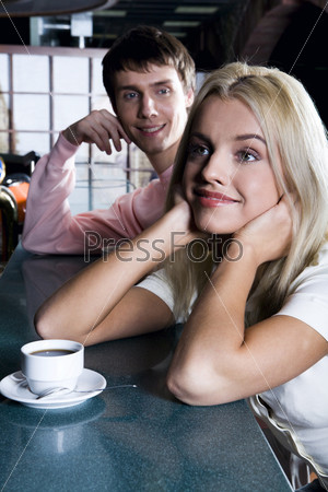 Smiling cute blond woman drinking coffee in the bar and handsome brunette man behind admiring her
