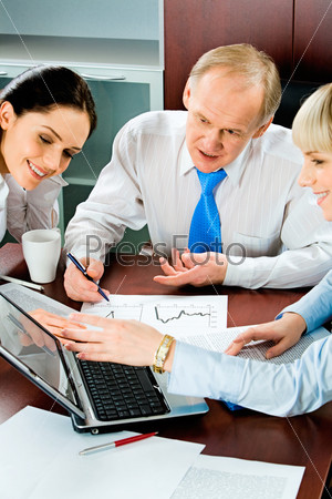 Vertical image of three business people sitting at the table and looking into the laptop monitor with one of businesswomen pointing at it with smile with some papers, pen and cup near by