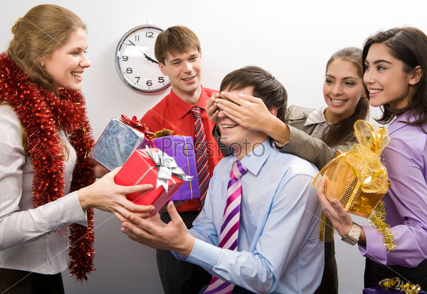 Photo of cheerful businesswoman keeping her hands on man?s eyes while their colleagues preparing gifts for him