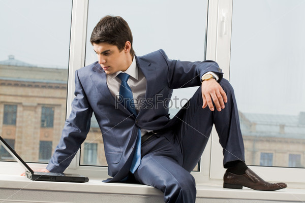 Photo of smart businessman sitting on window-sill with laptop near by and looking at monitor