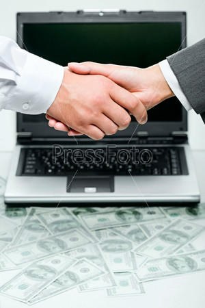 Business person shaking hands over opened laptop and heaps of money on the background