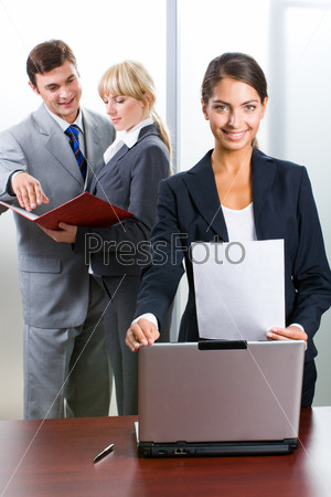 Young successful woman is standing near her co-workers and looking at camera in the office