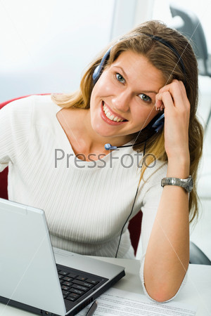 Portrait of beautiful smiling blond businesswoman in white blouse holding a pen in her hand and touching headset staring at the camera