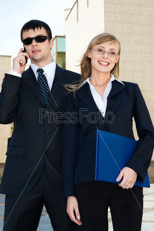 Beautiful smiling blond woman holding document-case in her hands and big man in sunglasses talking by the cell phone behind her