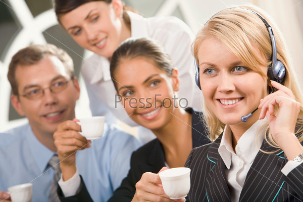 Portrait of cute blond woman with headset and her three colleagues behind gazing at her holding cups