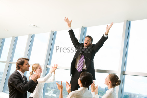Photo of successful businessman raising his arms and shouting surrounded by his colleagues applauding