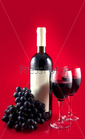 Two glasses with dark red wine on a red background. A dark wine bottle with a white label. Red ripe grape.