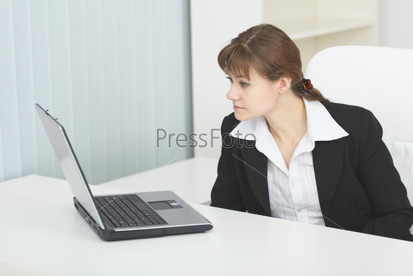 The young serious woman sits at a table at light office with the laptop
