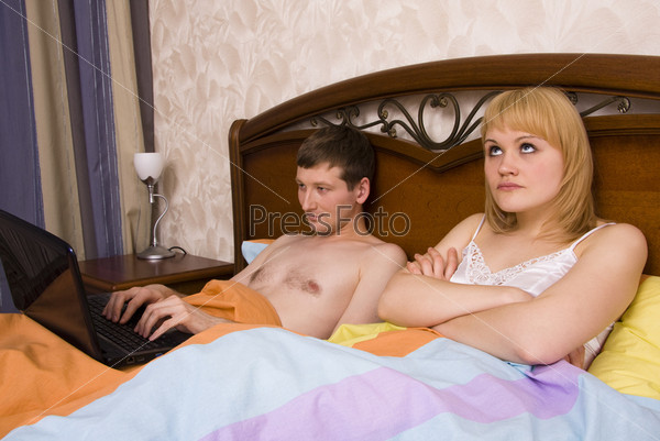 Young couple in a bed and bored woman. Male is working on computer and female with sad expressions. Woman and man having a disagreement.