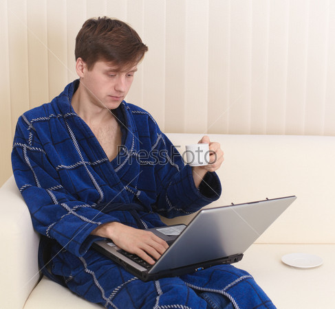 Мужчина в домашнем халате пьет кофе и смотрит в ноутбук, сидя на диване