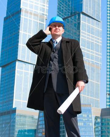 Businessman in hard hat talking on cell phone and holding drawing in his hand,on skyscrapers background