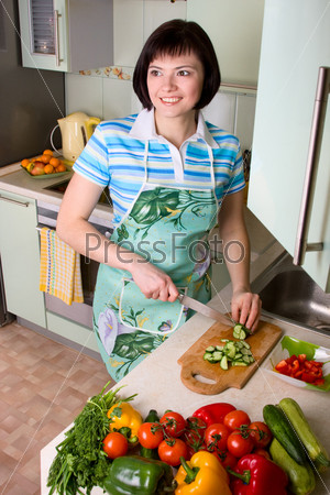 Young woman cutting vegetables in the kitchen.  Pretty girl in is cooking in the kitchen. home life: woman preparing something to eat. Smiling female standing and cutting cucumber in the kitchen.