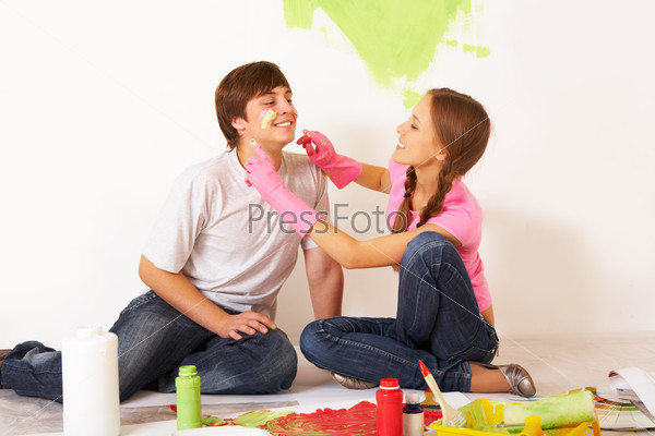 Image of joyful girl painting lad?s face with green color while having fun during improvement of flat