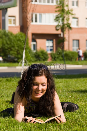 High school student, lying in grass on school campus reading a book. Student studying on the grass. Beautiful young woman reading book at park