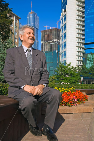 smiling Senior man in suit sitting in park near skyscrapers