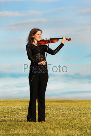 girl stands on grass and plays violin against  sky