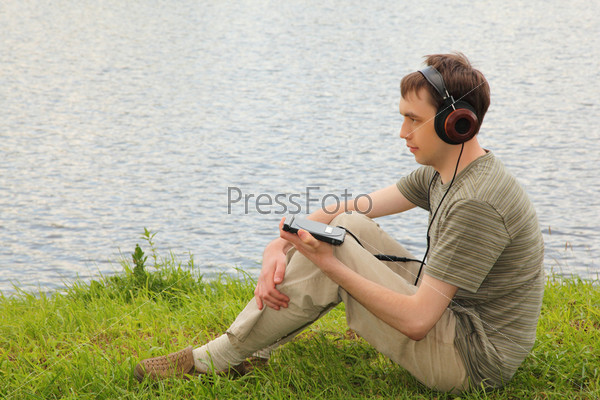 Young man listens music in headphones sits on grass ashore