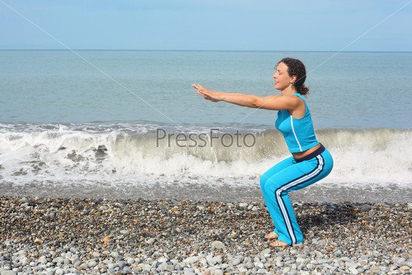 joyful woman wearing sporty clothes is making exercise on sea coast. she is squating.