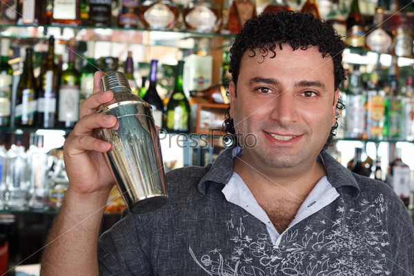 Barman with shaker behind bar rack. Smiling man against shelves with bottles.