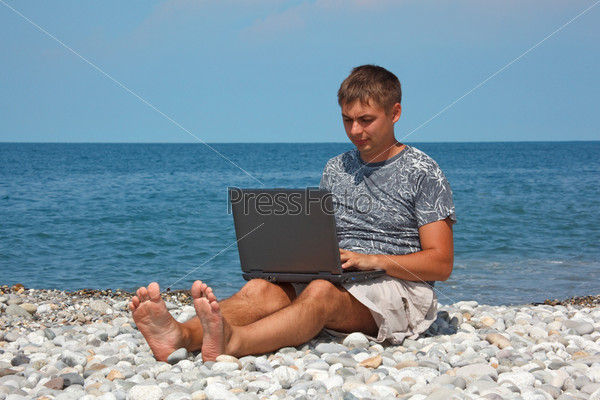 Man sitting on beach with laptop on his knees against  backdrop of sea.