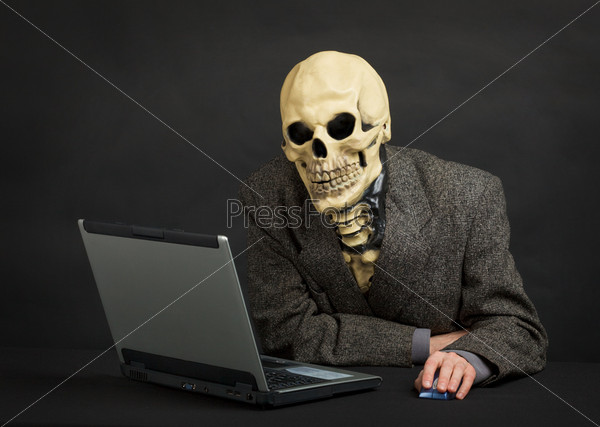The terrible skeleton in a suit sits at black office with the laptop