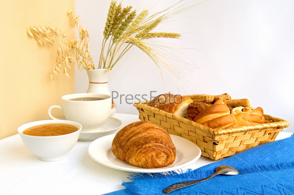 Table with a cup of tea and croissant on a plate, rolls in a wicker basket, jam in a bowl, stalks of wheat, oats and rye in a vase, blue cloth, a teaspoon against a background of white and beige curtains, stock photo