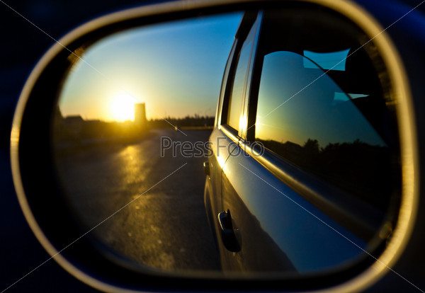 A sunset in the rearview mirror of car as a races down the road. Window of car is opened
