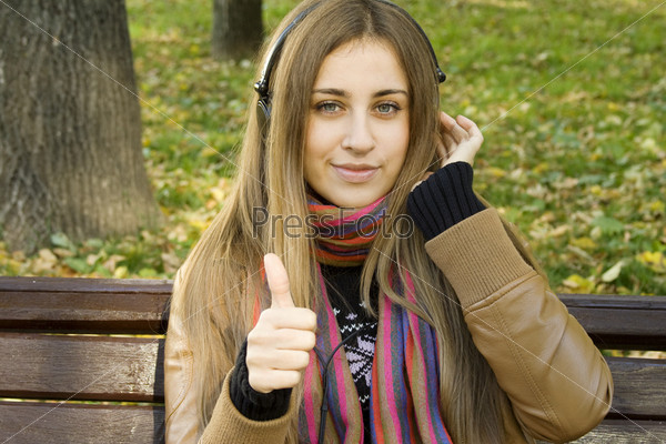 Young Caucasian woman with headphones in autumn park sitting on a wooden bench, listening to music. Thumbs up. Autumn around a lot of colorful foliage