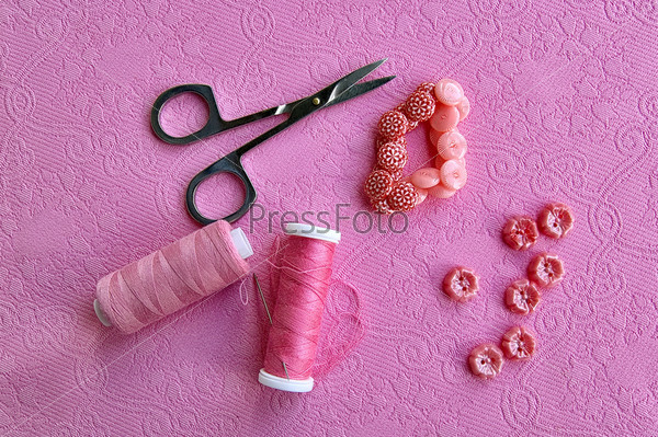 Pink spool of thread, needles, scissors, pink buttons on the background of a pink patterned fabric, stock photo