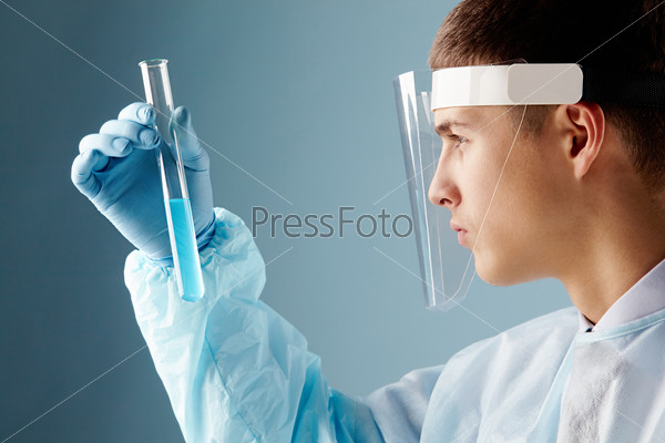 Serious clinician looking at flask with blue liquid through transparent safe mask, stock photo