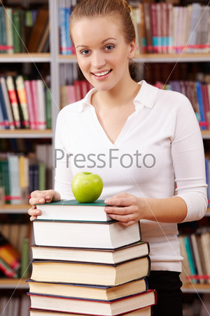 Portrait of pretty female looking at camera with green apple on top of book heap near by