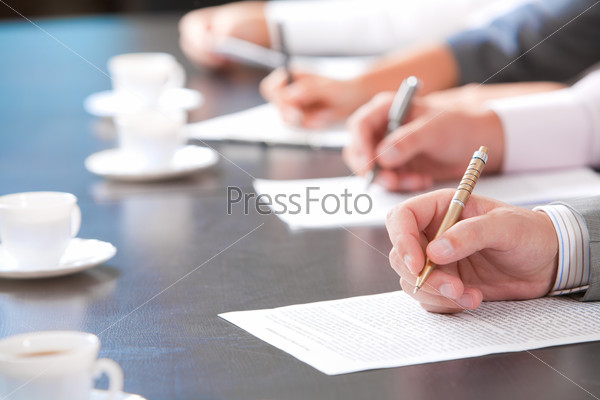 Close-up of masculine hand holding ballpoint over business document on background of human hands with cups of coffee near by