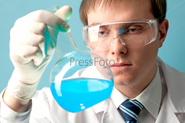 Serious clinician looking at flask with blue liquid in laboratory, stock photo