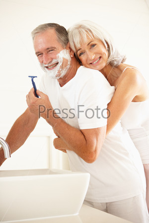 Senior Man Shaving In Bathroom Mirror With Wife Watching