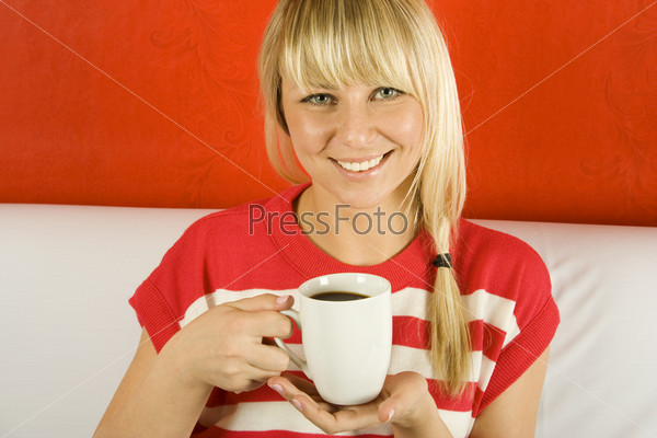 Beautiful young woman is drinking coffee from a white mug sitting on the couch