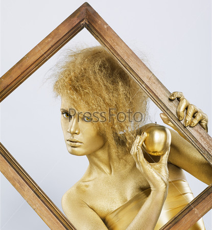portrait of girl with golden bodyart posing with golden apple in her hands in picture frame on gray