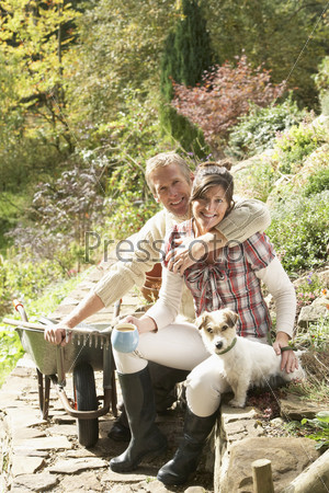 Couple With Dog Having Coffee Break Whilst Working Outdoors In Garden