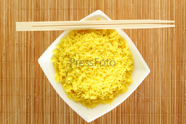 Boiled yellow rice and a white plate beside lie wooden sticks. Plate stands on a wooden mat