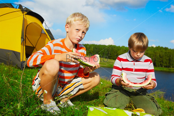 outdoor portrait of two boys eating watermelon on green grass near camp