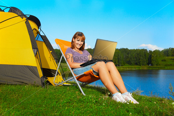 outdoor portrait of beautiful happy woman with laptop sitting in folding chair near camp tent