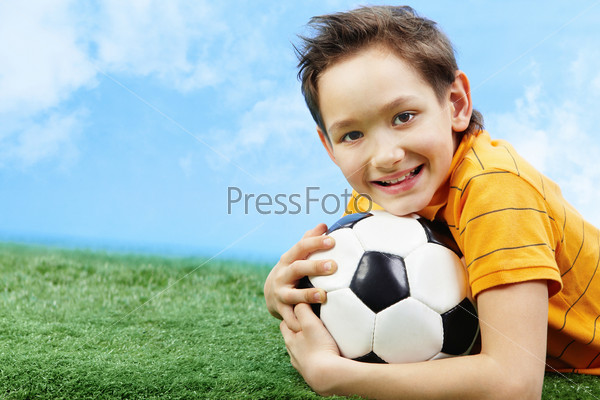 Image of happy boy lying on the grass with ball and looking at camera